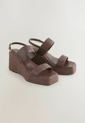 Bruine wedge sandalen met twee verstelbare banden en een gevlochten touwzool, geplaatst op een lichte achtergrond.