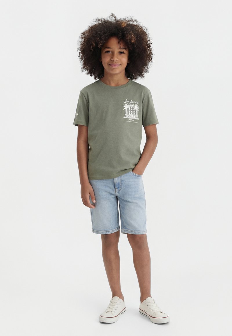 Barn med krøllet hår iført en grøn t-shirt med grafik, lyse denimshorts og hvide sneakers, stående med den ene hånd i lommen.