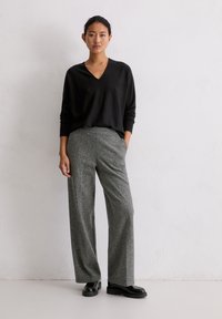 Schwarzer V-Ausschnitt-Pullover kombiniert mit grauen gemusterten weit geschnittenen Hosen und schwarzen Schuhen. Das Outfit zeichnet sich durch schlichte Texturen und einen lockeren Schnitt aus.