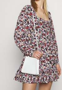 Abito floreale con maniche lunghe, scollatura quadrata e bordo arricciato. Borsa a tracolla bianca con finitura testurizzata. Sfondo scuro che contrasta con la stampa floreale.