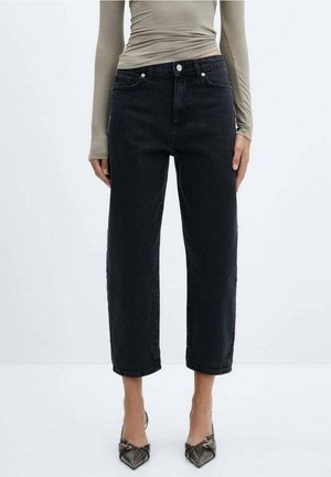 Femme portant un jean noir taille haute, droit et court, un haut beige à manches longues et des escarpins noirs à bride arrière pointus, debout sur un sol blanc uni.