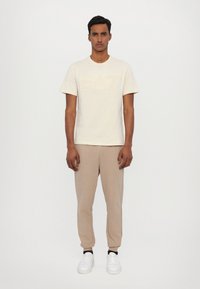 Lichtbeige t-shirt met een subtiele reliëf EA7-logo. Gecombineerd met beige joggingbroeken en witte sneakers, wat een gecoördineerde, casual look creëert.