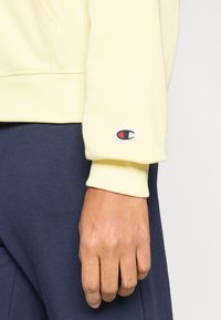 Ανοιχτό κίτρινο φούτερ με μανίκια με μανσέτες που διαθέτουν μικρό κεντημένο λογότυπο σε κόκκινο και μπλε. Λείο ύφασμα, φοράει πάνω από σκούρο παντελόνι.