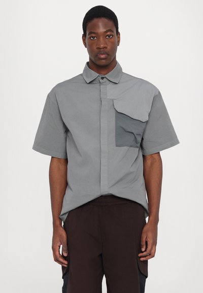 Chemise grise à manches courtes avec un devant à boutons, présentant un détail de poche asymétrique et une texture lisse, assortie à un pantalon sombre.