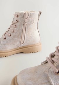 Stivaletti alla caviglia rosa chiaro con superficie texture, chiusura con lacci sul davanti, zip laterale e suola in gomma. Fodera in pile visibile nella parte superiore.