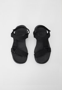 Mustat sandaalit, joissa on teksturoitu kumipohja ja säädettävät kangasvyöt, sekä yksinkertainen muotoilu ja avoin kärki.