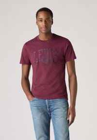 Burgunderfarbene Baumwoll-T-Shirt mit dem "LEVI'S"-Logo in dunklerem Farbton, runder Ausschnitt, kurze Ärmel, kombiniert mit hellblauen Jeans.