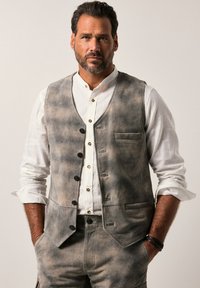 Chaleco gris y beige con un acabado texturizado, con un escote en V, seis botones negros y bolsillos laterales, llevado sobre una camisa blanca.