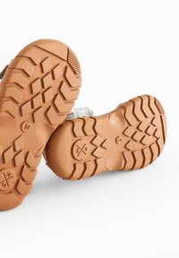 Brun gummisål med dybe mønstre. Lys grå overdel med stropper og teksturerede detaljer. Designet til godt greb og holdbarhed.