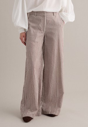 Frau trägt hoch taillierte, weit geschnittene braune Hose mit weißen vertikalen Streifen und eine weiße Langarmbluse, steht mit den Händen in den Taschen.