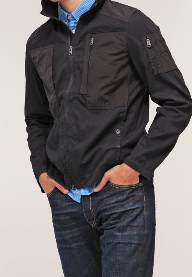 Giacca nera con zip e molteplici tasche, caratterizzata da pannelli opachi e lucidi a contrasto. Indossata sopra una camicia a quadri blu e jeans scuri.