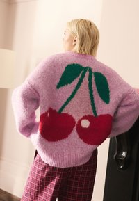 Ružový chlpatý sveter s grafickým motívom čerešne v červenej a zelenej farbe na chrbte. Rebrovaný lem a široké rukávy pre pohodlný strih.