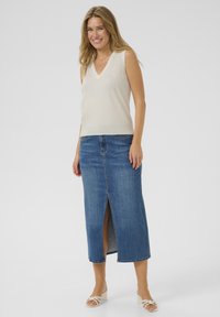 Top sans manches en V de couleur beige clair, associé à une jupe en denim bleue taille haute avec une fente à l'avant et des bords ourlés. Sandales à talons blocs blanches.