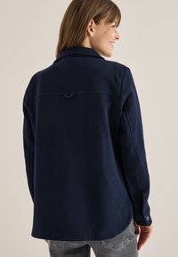 Chaqueta azul marino con un acabado texturizado, que presenta un collar, puños abotonados y un detalle de lazo en la espalda. Ideal para superponer.