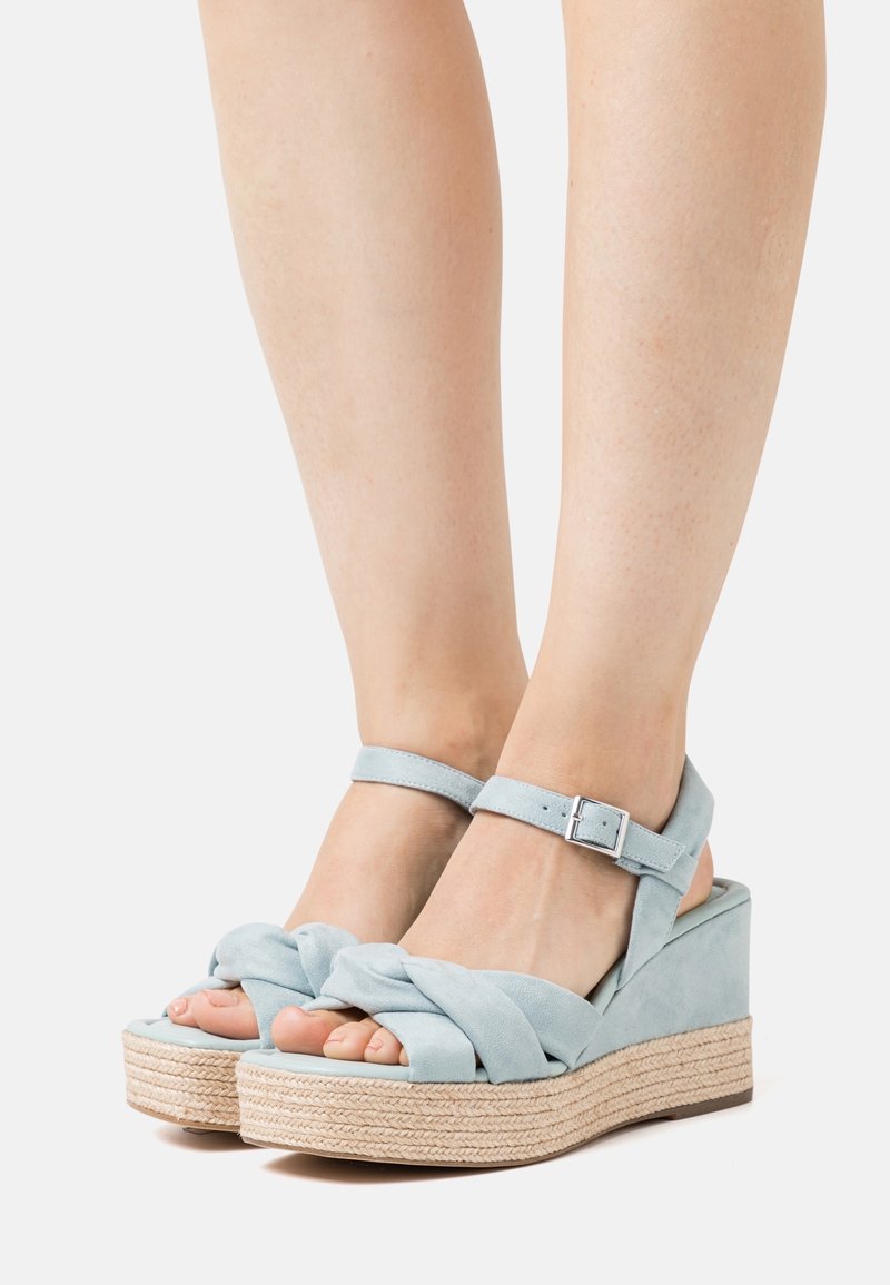 Sandales compensées en daim bleu clair avec brides à la cheville et design noué à l'avant, portées pieds nus, montrant des semelles plateforme tissées texturées.
