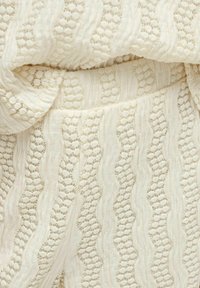 Tela de color crema con patrones texturizados que presentan líneas onduladas y acentos circulares. Material suave y ligero con un acabado delicado.