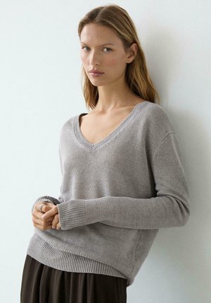 Jeune femme aux longs cheveux blonds portant un pull gris clair à col en V et un pantalon foncé, debout contre un mur blanc les mains jointes.