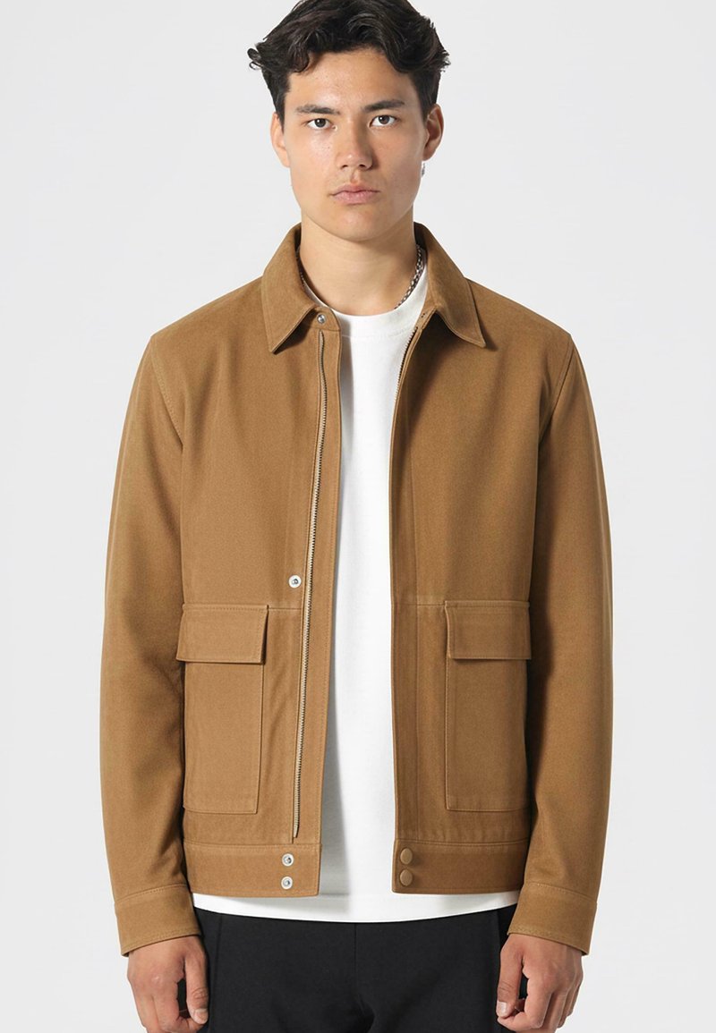 Brązowa, teksturowana kurtka z kołnierzykiem, z przodu na zamek błyskawiczny, dwoma przednimi kieszeniami oraz zapięciami na napy na dole. Nosi się ją na białej koszuli.