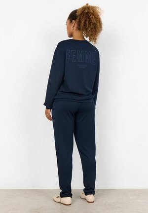 Felpa blu navy e pantaloni abbinati realizzati in morbido tessuto, con dettagli testuali "FEMME" sul retro. Indossati con calzature chiare.