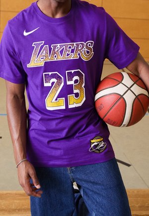 Person hält einen Basketball, trägt ein lila Lakers-T-Shirt mit der Nummer 23 und blaue Jeans, steht drinnen auf einem Holzboden.