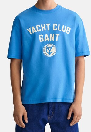 Camiseta de algodón azul con mangas cortas, que presenta "YACHT CLUB" y "GANT" impresos en blanco sobre un diseño de logo en forma de círculo.