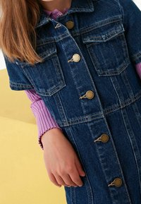 Denim-mekko, jossa on kaulus, isot etutaskut ja kullatut napit. Ominaisuuksiin kuuluu tummansininen väri ja kontrastiväriset tikkaukset. Päällä on purppuranvärinen ribbimäinen paita.