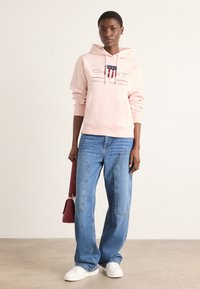 Lichtroze hoodie met bedrukte logo, gecombineerd met wijde blauwe denim jeans en witte sneakers. Houdt een kleine rode handtas vast.
