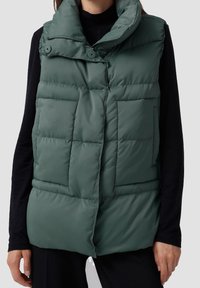 Grüne gepolsterte Weste mit hohem Kragen, ausgestattet mit zwei frontalen Taschen und einem Druckknopfverschluss. Glatte Textur und strukturierte Form.