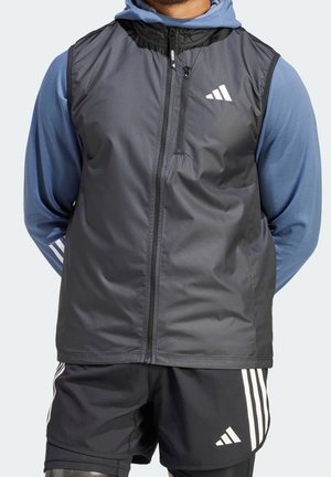Mężczyzna w czarnej bezrękawniku, pod którą ma niebieską bluzę z kapturem oraz czarne spodenki Adidas z białymi paskami na neutralnym tle.
