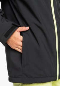 Čierna nepremokavá bunda so svetlozeleným zipsom a suchým zipsom na manžetách. Obsahuje bočné vrecká a hladkú, trochu texturovanú tkaninu.