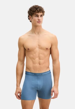 Hemdloser Mann mit lockigem Haar, der blaue Marc O'Polo Boxershorts trägt und vor einem schlichten hellen Hintergrund steht.