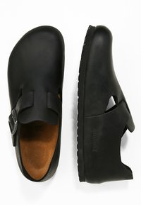 Fekete klumpák nyitott orr kialakítással, bőr felsőrésszel, állítható csattal és gumitalppal, amely texturált részletezéssel rendelkezik.