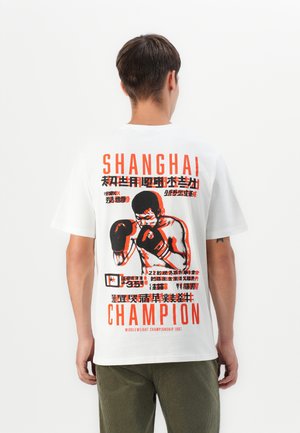 T-shirt en coton blanc avec un graphique audacieux en rouge et noir d'un champion de boxe, avec le texte "SHANGHAI" et "CHAMPION 1987" au dos.