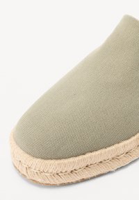 Chaussure slip-on en toile verte avec un bout arrondi et une bordure en jute de style espadrille. Surface texturée et détails en fibres naturelles.