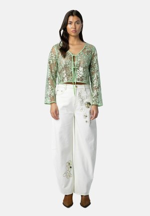 Kobieta w przezroczystej zielonej bluzce z długim rękawem z kwiatowej koronki, zawiązanej z przodu, białe szerokie spodnie z kwiatową haftem i brązowe, szpiczaste buty.