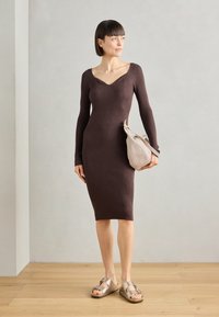 Vestido de punto, de manga larga, color marrón chocolate, con una forma ajustada y textura acanalada. Combinado con un bolso beige claro y sandalias marrones.