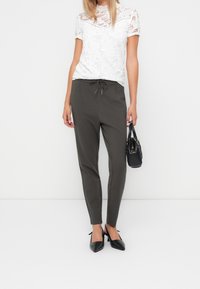 Donna che indossa un top bianco in pizzo a maniche corte, pantaloni grigi a sigaretta con cordoncino, tacchi slingback neri e tiene in mano una borsa nera.