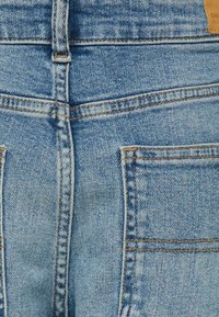 Svijetloplave traperice s visokim strukom. Sadržavaju duple stražnje džepove, vidljivo šivanje i kožnu oznaku branda na struku.