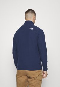 Marine fleecejakke med høj krave, med et logo på den øverste del af ryggen, og en glat tekstur. Ærmekanter med justerbare stropper.