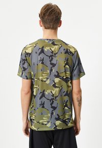 Camisa de manga corta con patrón de camuflaje, con una textura de malla transpirable en tonos verdes, grises y negros. Cuello redondo, corte relajado.