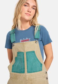 Pettorina beige con tasche a contrasto color teal, accessori in metallo e spalline regolabili, abbinata a una maglietta blu a costine con stampa grafica.