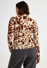 Donna con capelli corti scuri che indossa una maglietta a maniche lunghe beige e marrone a pois e pantaloni grigio scuro, in piedi contro uno sfondo bianco uniforme.