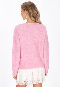 Flauschiger rosa Pullover mit rundem Halsauschnitt und langen Ärmeln, ausgestattet mit gerippten Bündchen und Saum. Über einem weißen Spitzenkleid getragen.