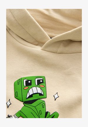 Felpa con cappuccio beige con un personaggio dei cartoni animati verde dalla testa quadrata e un'espressione arrabbiata. Il tessuto ha una texture liscia e cuciture visibili.