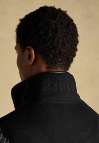 Melnas vilnas veste ar augstu apkakli, ar izsistām "JOULES" logotipu. Tekstūrēta auduma un bezšuvju dizains redzams no aizmugures.