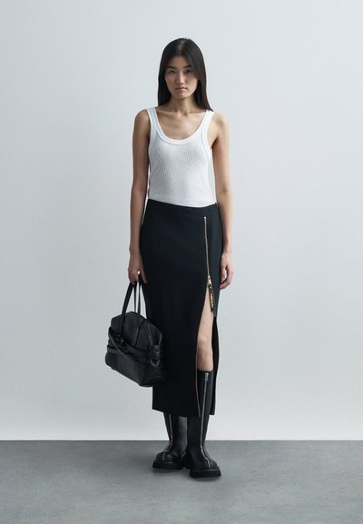 Femme portant un haut sans manches blanc, une jupe midi noire avec une fente à zip à l'avant, des bottes noires, tenant un sac à main noir, debout sur un sol gris.