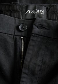 Gros plan sur un pantalon noir montrant un bouton, une fermeture éclair, un trou de bouton et une étiquette de ceinture avec le texte "aprel".