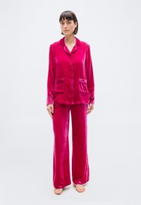 Γυναίκα που στέκεται ξυπόλητη φορόντας ένα φωτεινό ροζ βελούδινο σετ πυτζάμα με σακάκι με κουμπιά και παντελόνια φαρδιάς γραμμής, μπροστά σε απλό ανοιχτό φόντο.