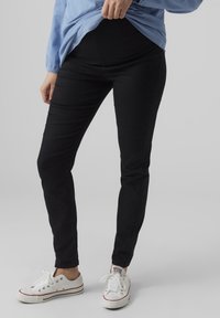 Crne slim-fit hlače od mekog materijala, s visokim strukom i glatkom teksturom, u kombinaciji s bijelim tenisicama i plavim topom.