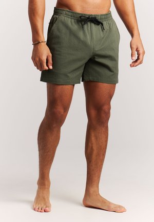 Man draagt olijfgroene zwemshorts met textuur en zwarte trekkoord, staat blootsvoets tegen een eenvoudige lichte achtergrond.